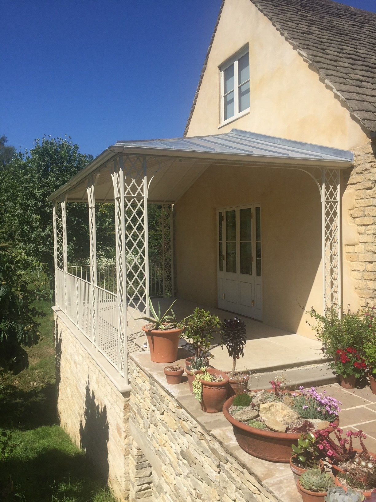 Beige house with porch, ornate white railing, potted plants, stone wall, blue sky.