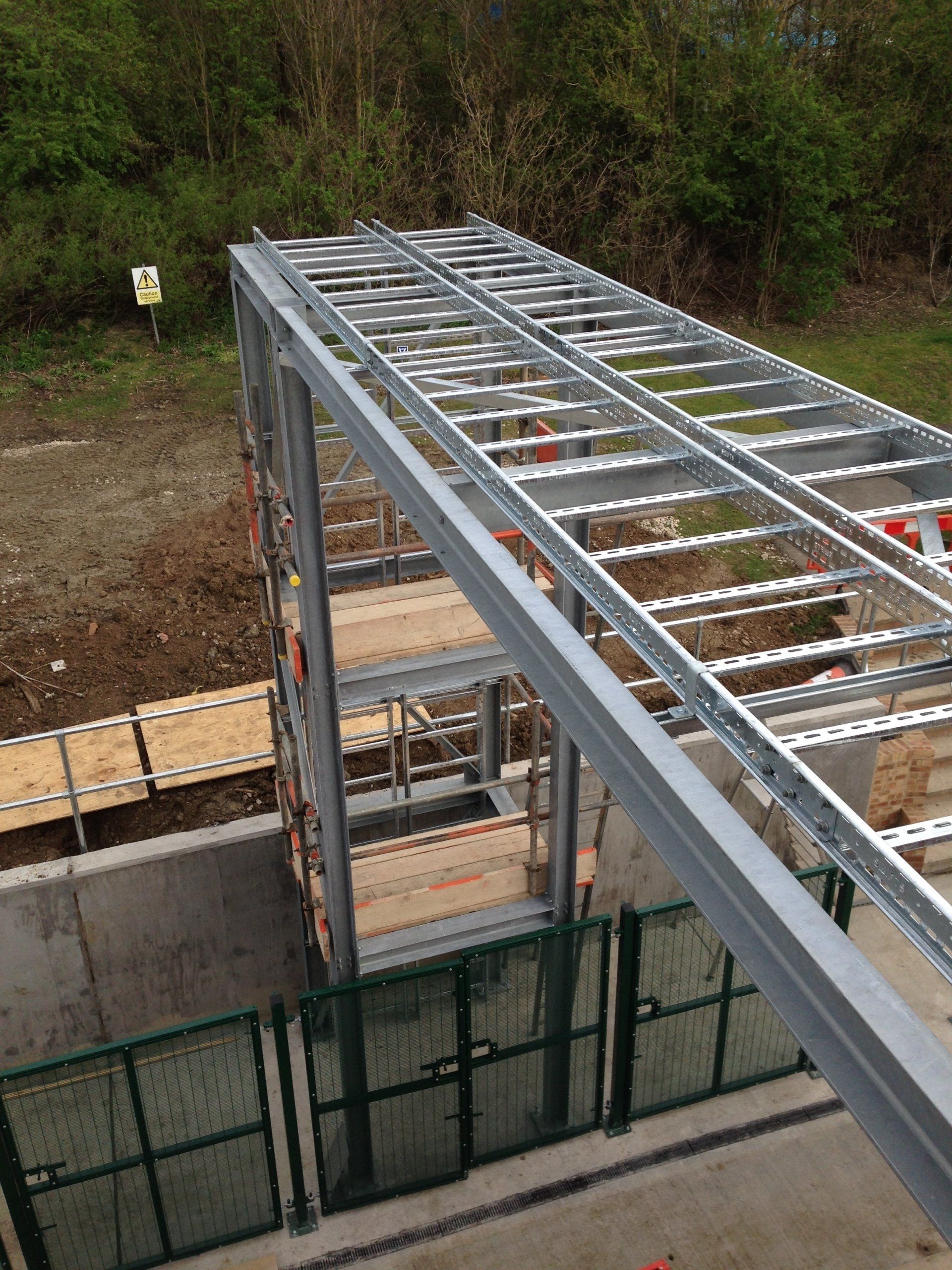 Steel framework structure, possibly a bridge or walkway, under construction, outdoors. Green fencing below.