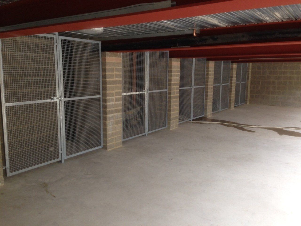 Metal mesh storage cages in a concrete room, attached to brick walls. Red beams and corrugated metal ceiling overhead.