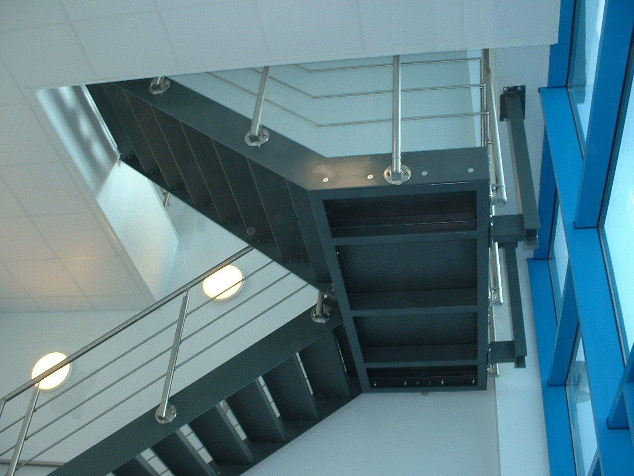 Gray metal staircase with stainless steel handrails against a white wall and blue window.