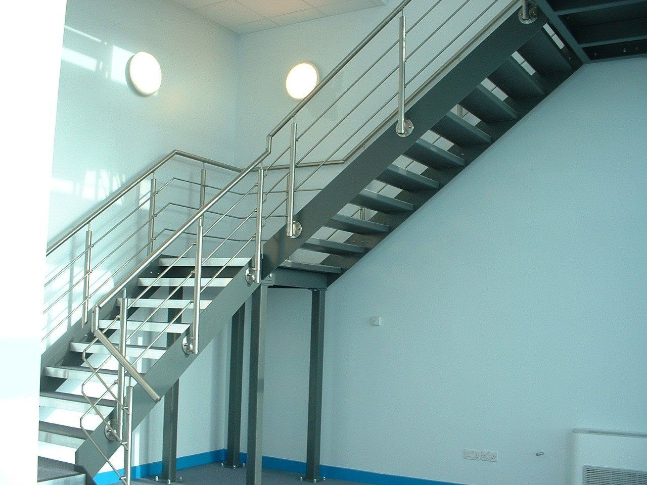 Metal staircase with railing ascending to the right. Light blue walls with two round lights.