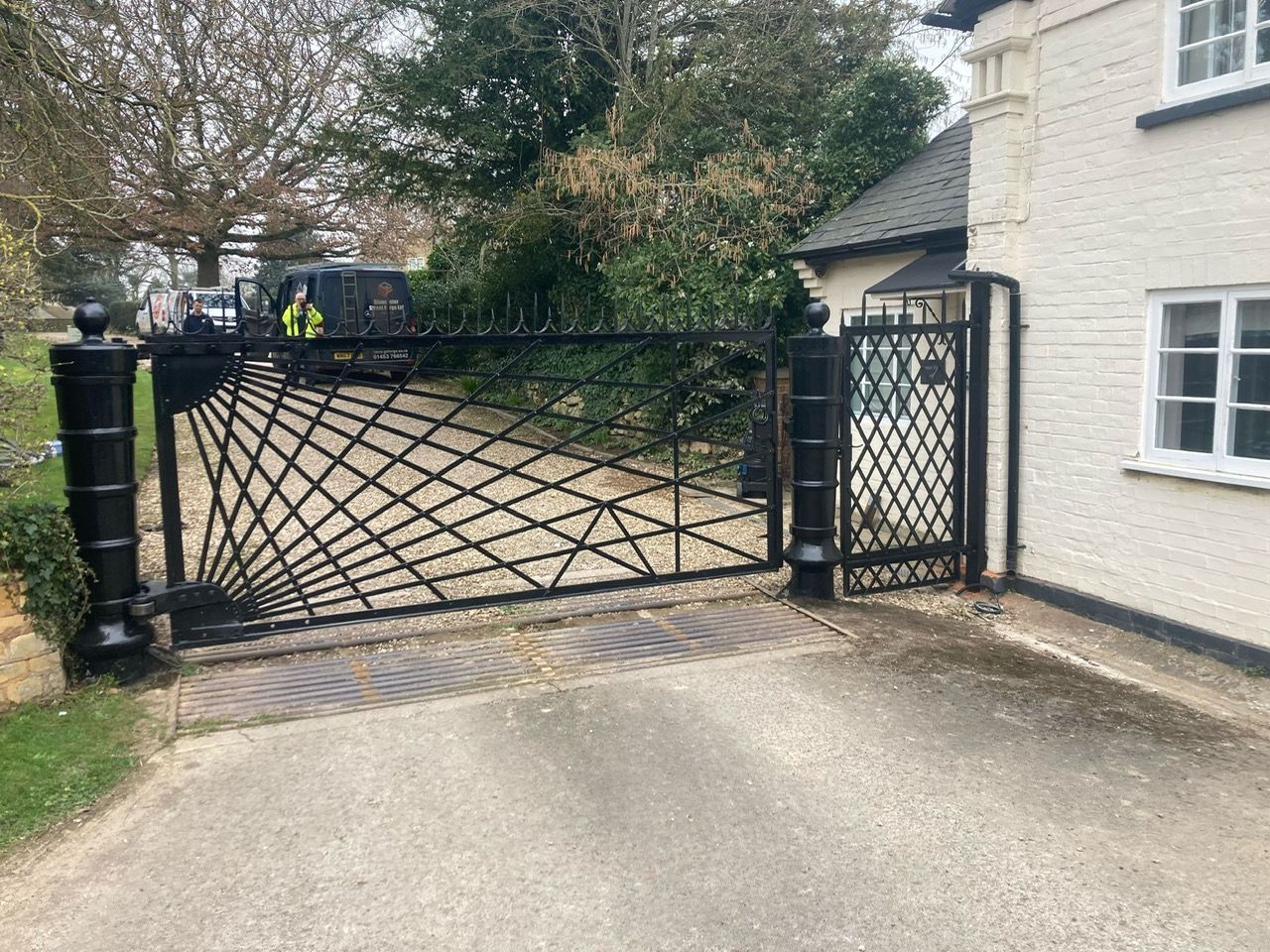 Black wrought iron gates with sunburst design, driveway leading to a white house with window.