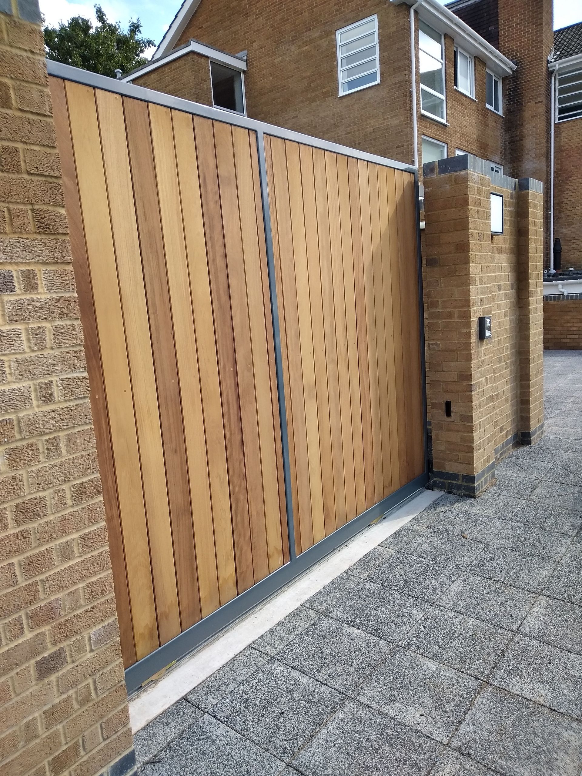 Wooden gate with vertical slats, set between brick pillars, leading to a driveway with a gravel surface.