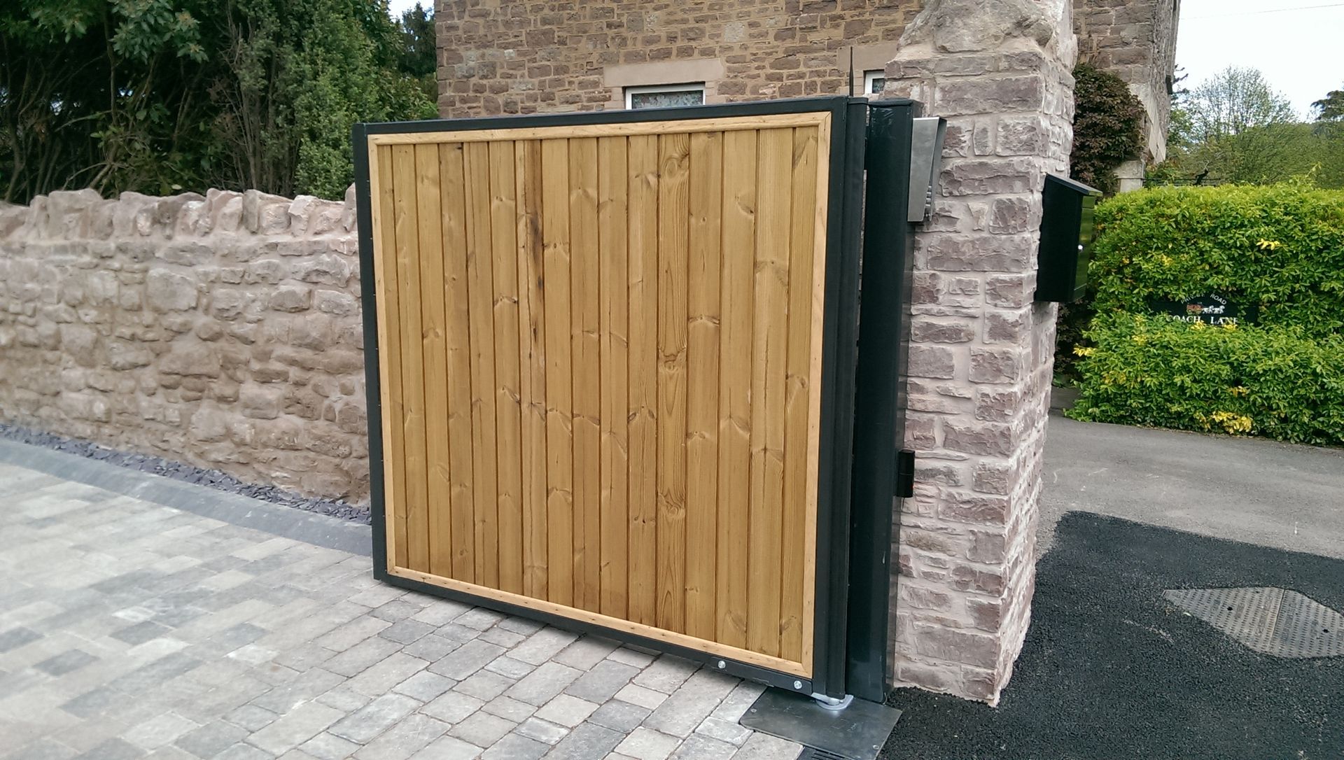 Wooden gate, tan vertical planks in black frame, on a paved driveway next to stone wall and brick structure.