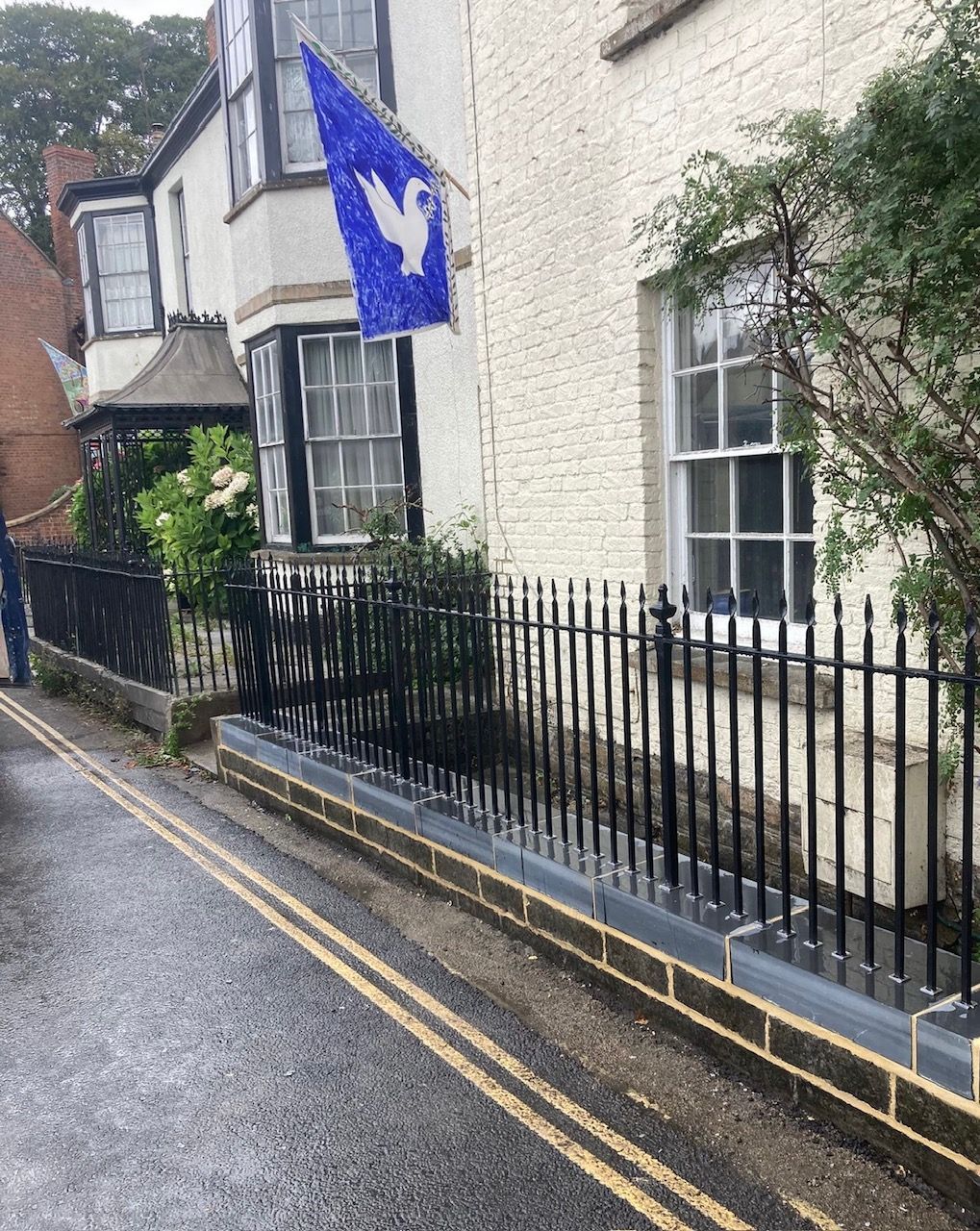 White building with blue flag. Black fence along a wet street with double yellow lines.
