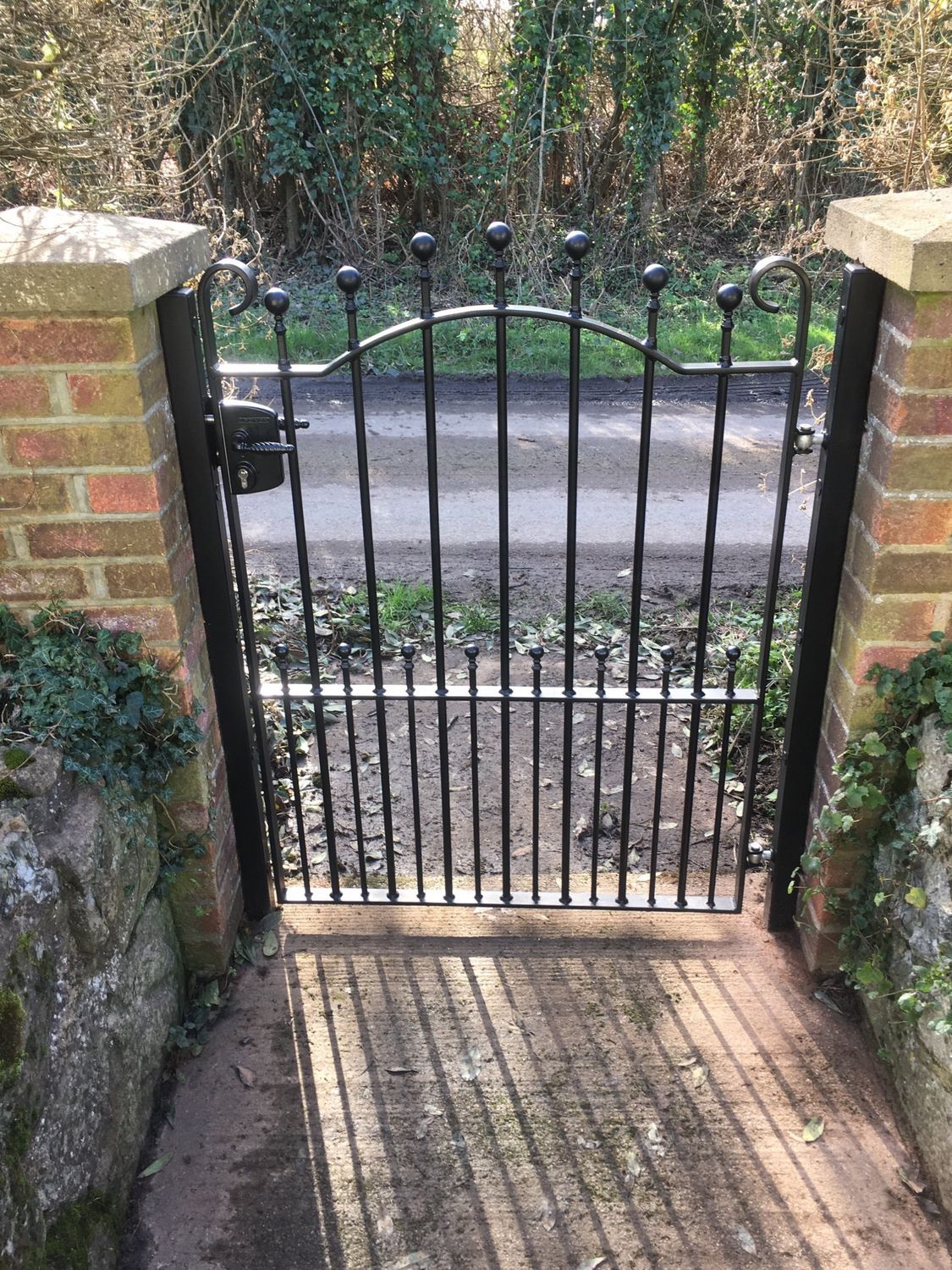 Black wrought iron gate set between brick pillars, with a concrete path leading up to it.