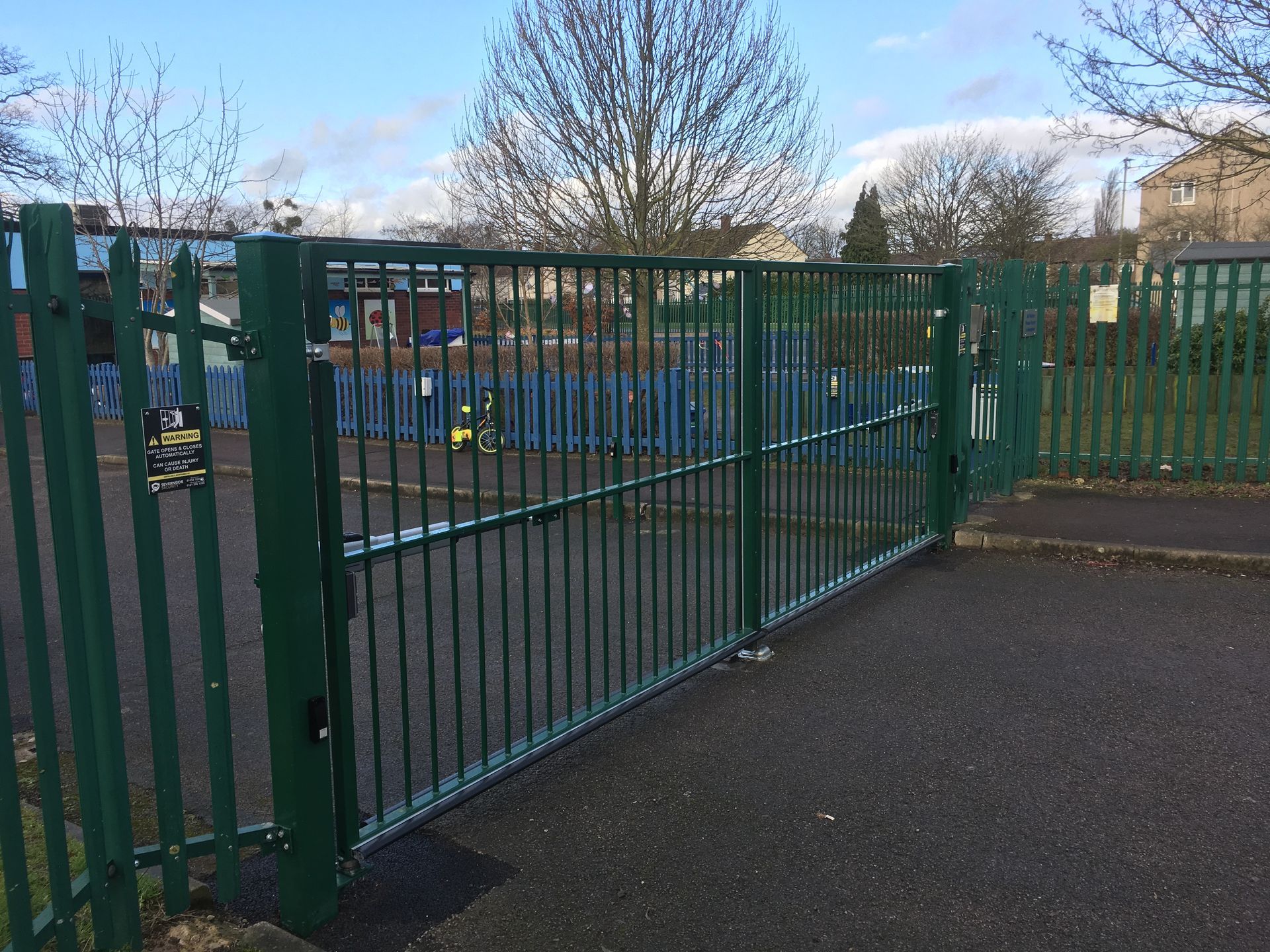 Green metal gate, closed, on asphalt. Fenced playground in background, trees and building visible.