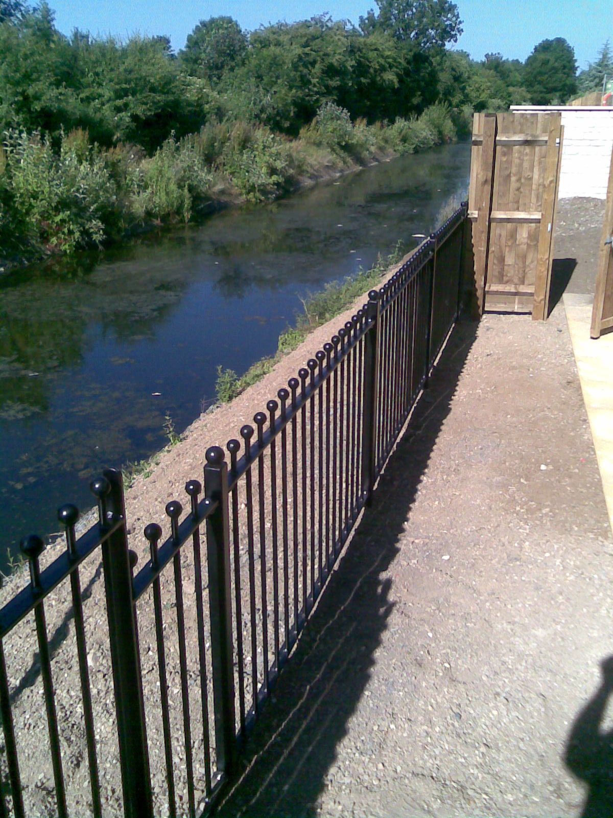 Black metal fence bordering a dark waterway with foliage in the background, under a blue sky.