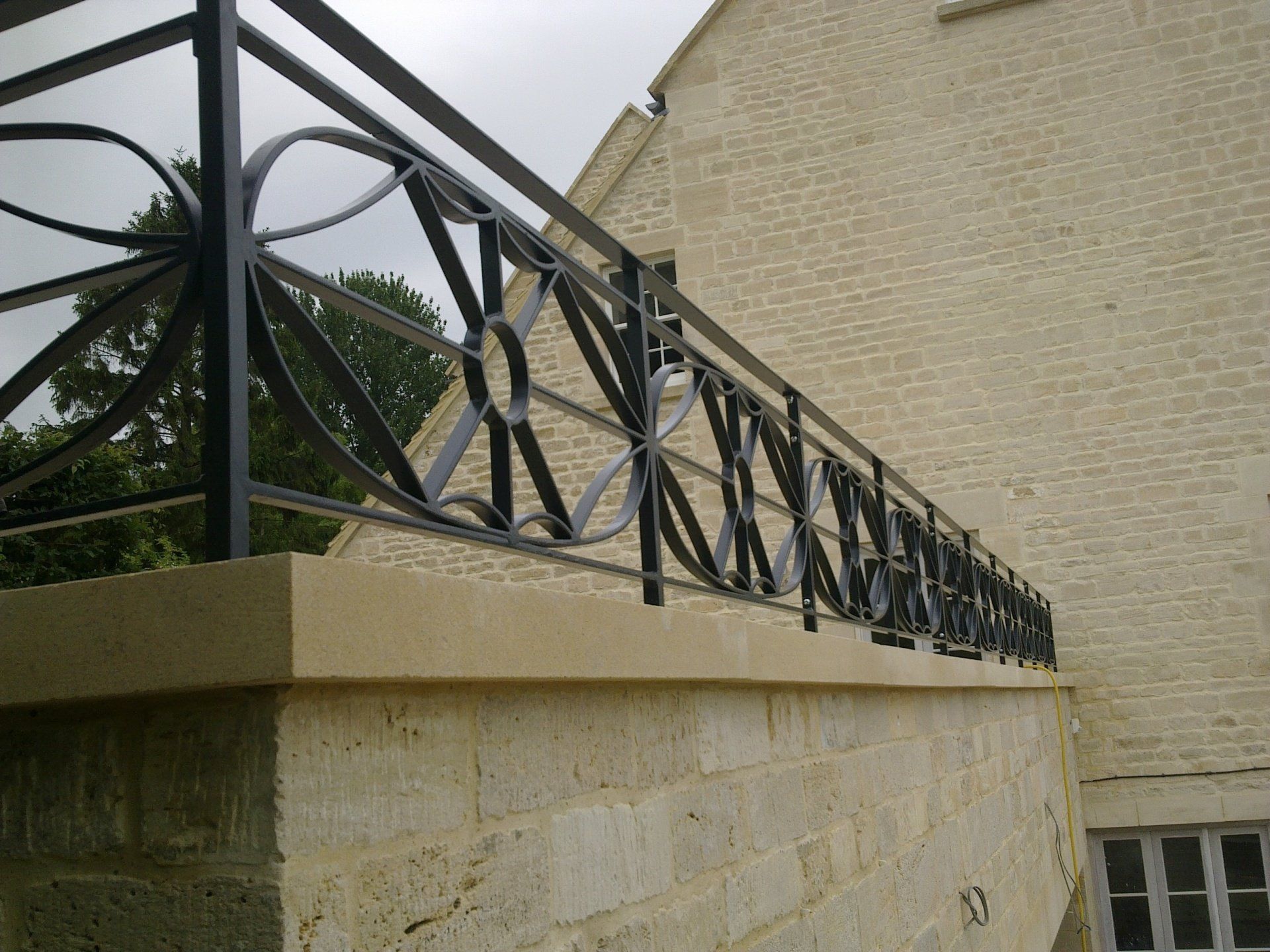 Black metal balcony railing on a light-colored stone wall. The railing has a floral design.