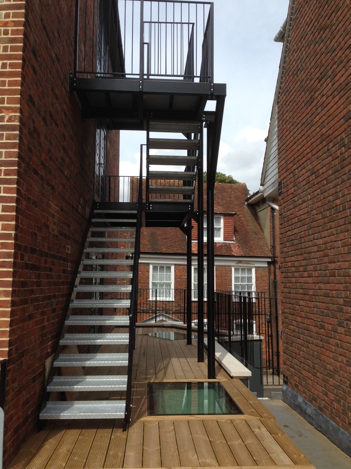 Metal staircase leading to a platform between brick buildings, with a small sunken wooden deck.