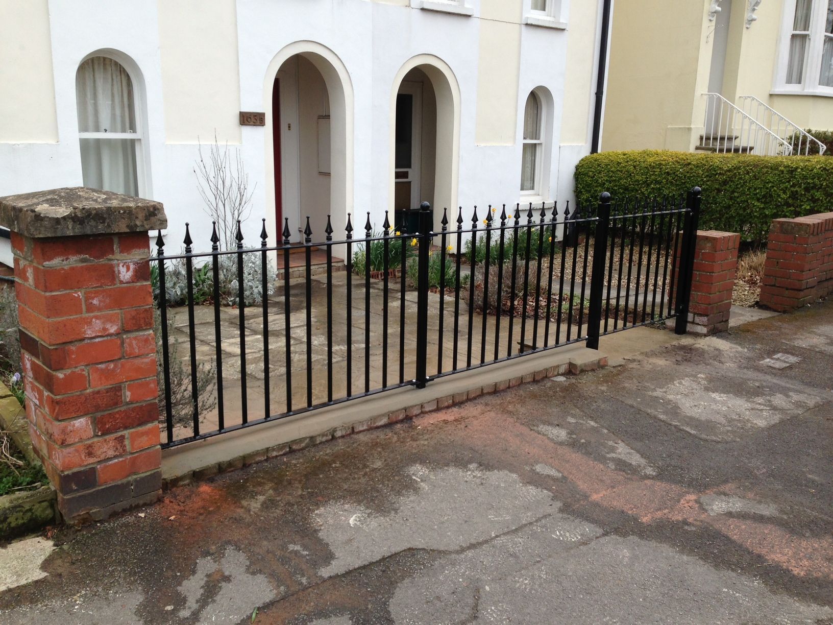 Black iron fence in front of a light-colored house with an arched doorway and windows, set on a brick foundation.