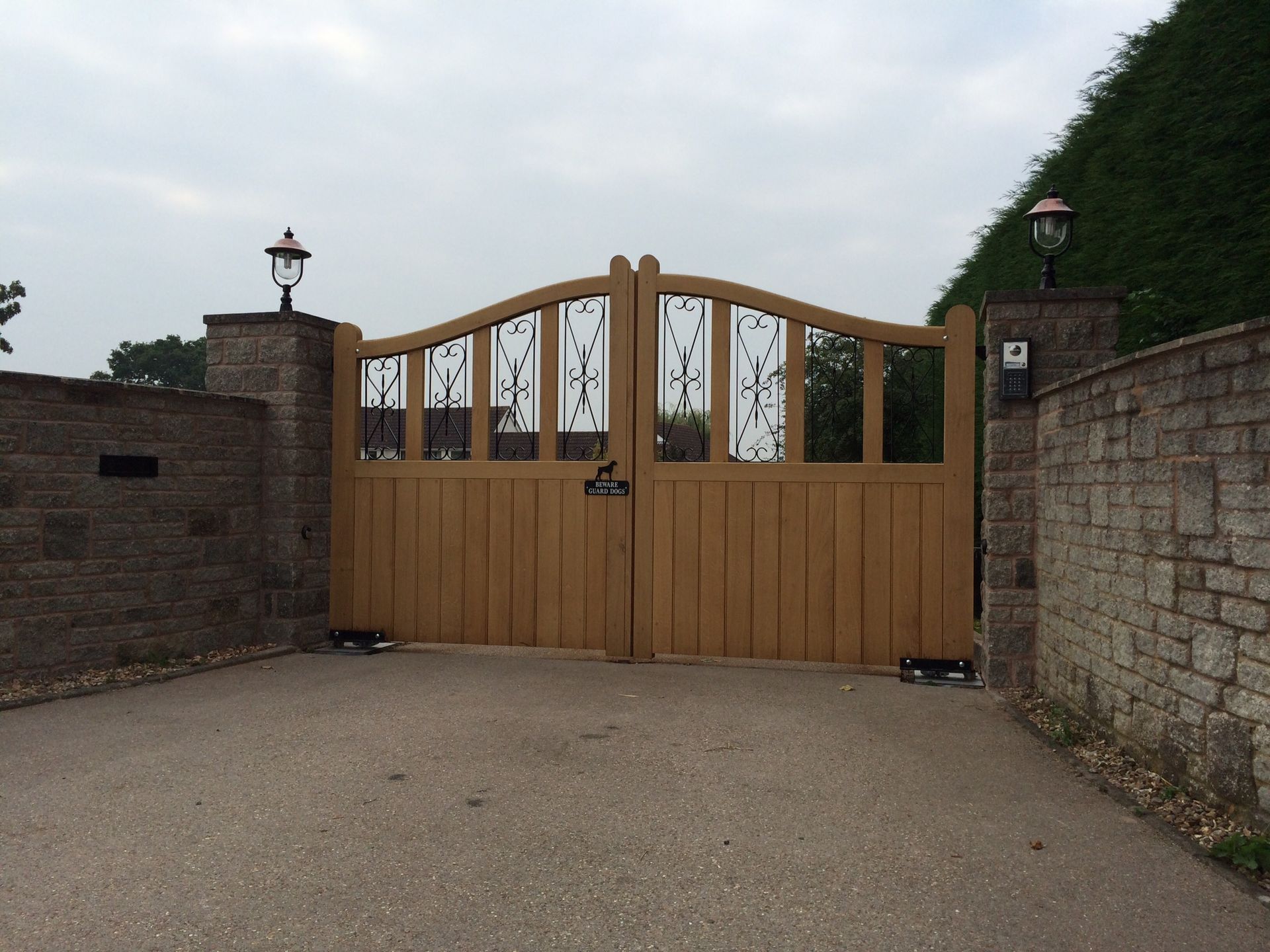 Wooden driveway gates with decorative ironwork, brick pillars, and sconce lights.