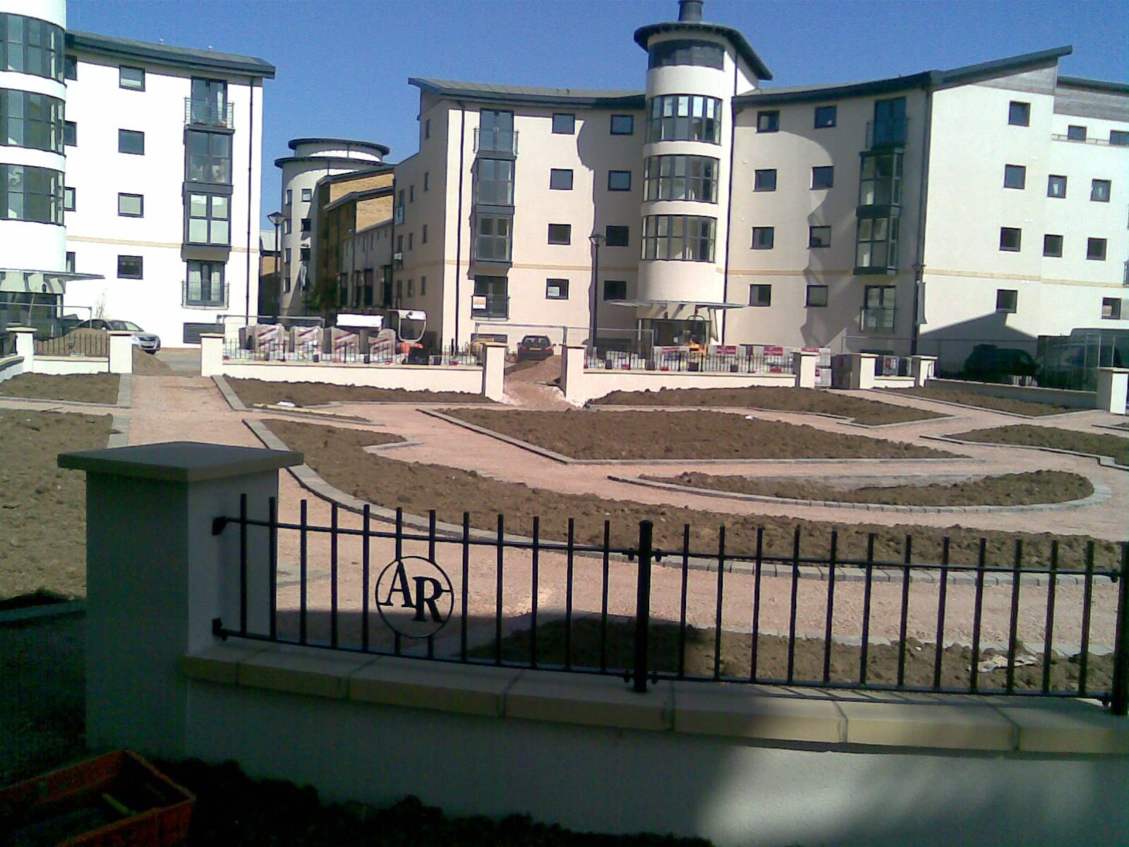 Exterior view of modern apartment buildings, with a small courtyard and a black metal fence in the foreground.