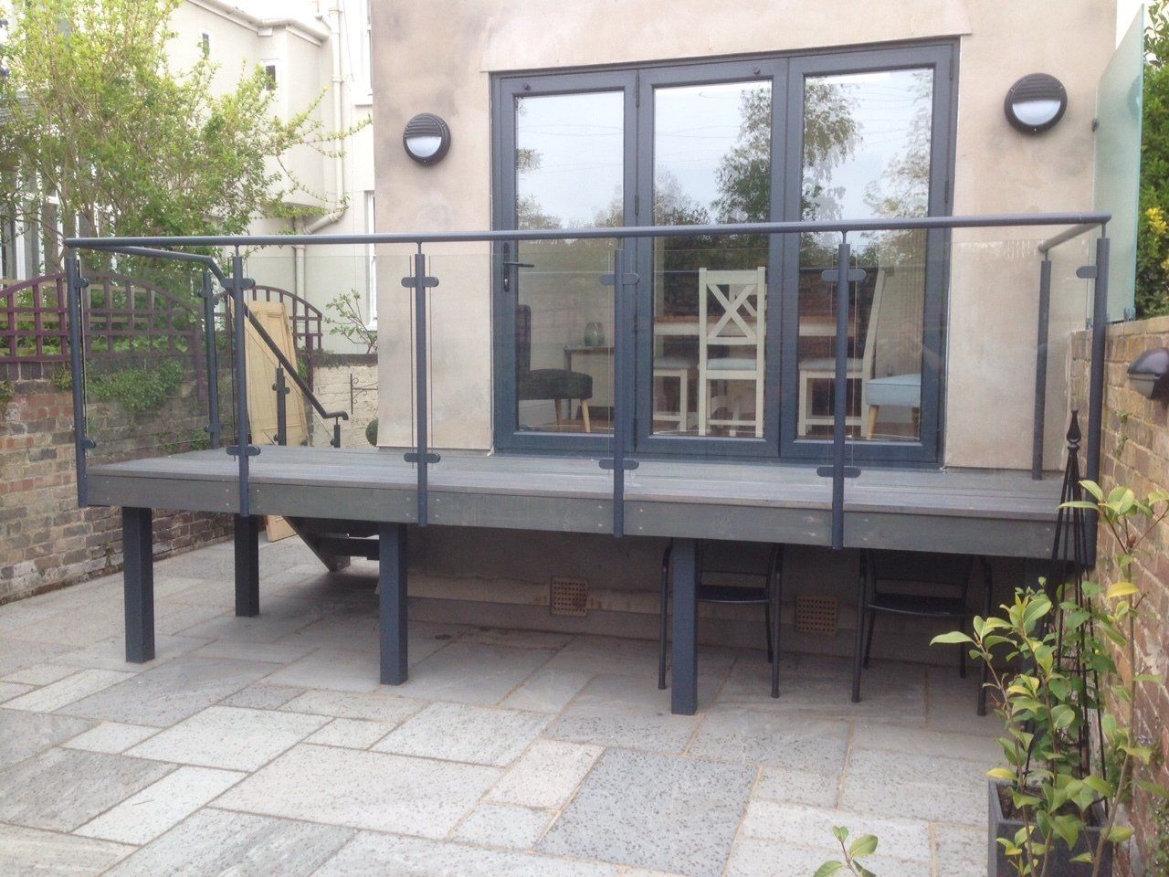 Grey deck with glass and metal railing, leading to double doors. Two round lights on the wall.