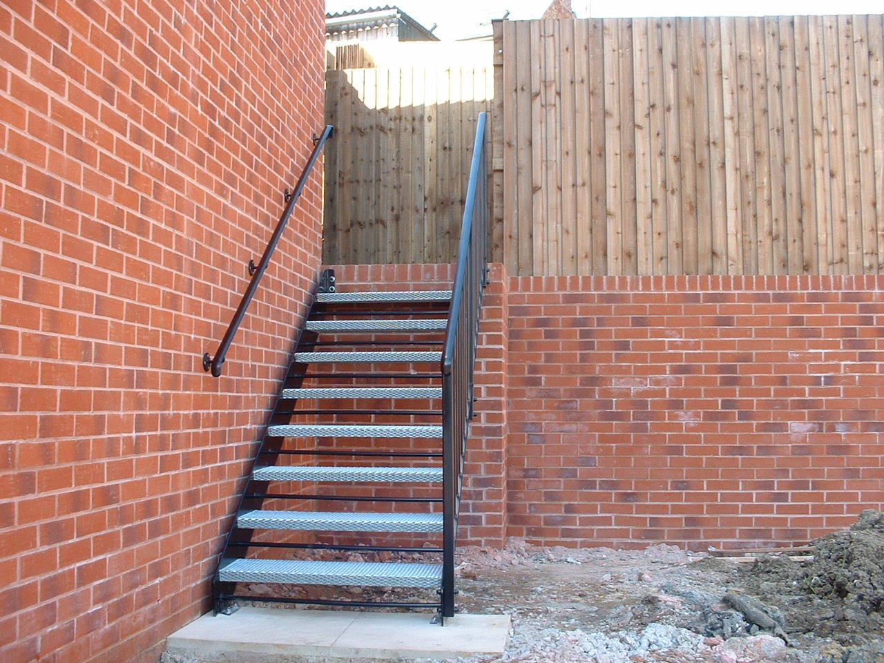 Metal staircase against brick wall with a wooden fence. Handrails present.