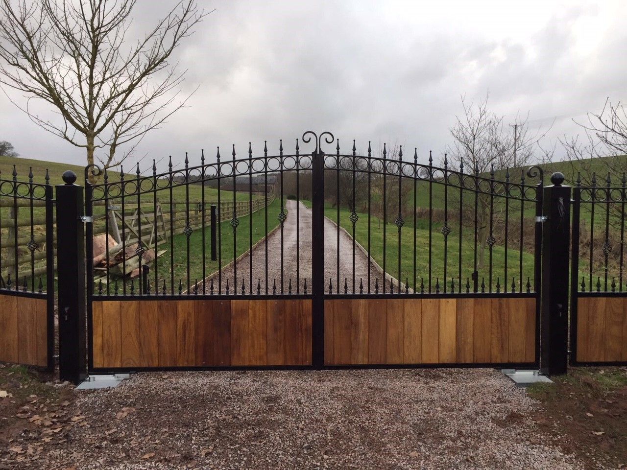 Black metal gates with arched top and wood paneling, opening to a gravel driveway. Cloudy sky.