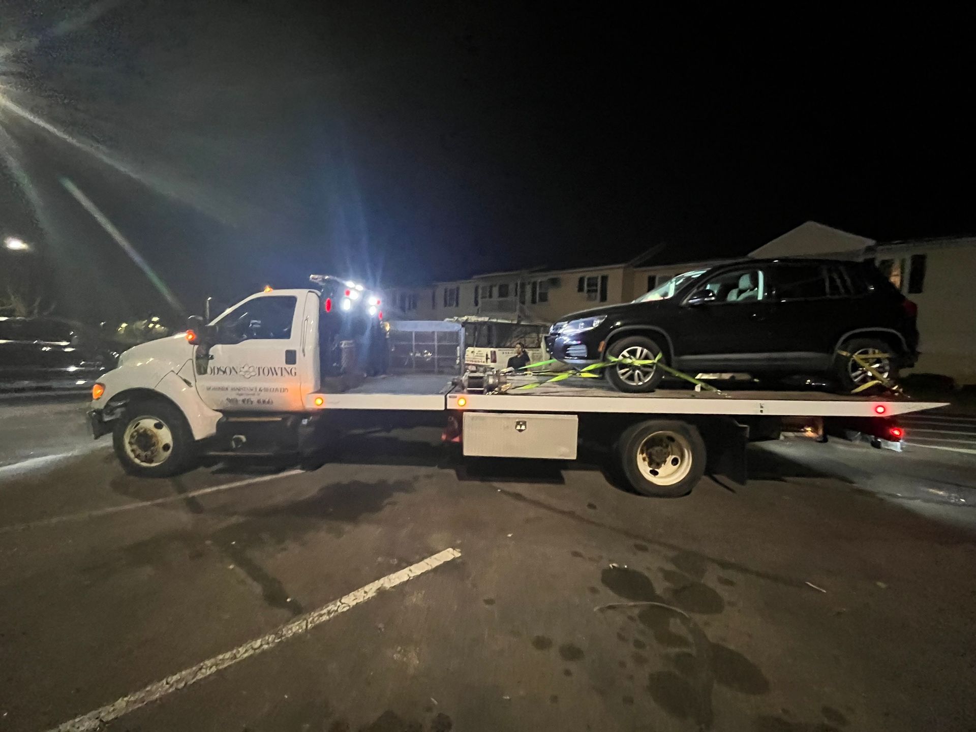 A tow truck is towing a black suv in a parking lot at night.