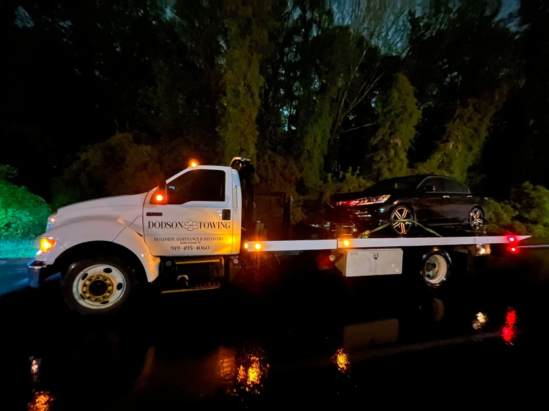 A tow truck with a car on the back is parked on the side of the road at night.
