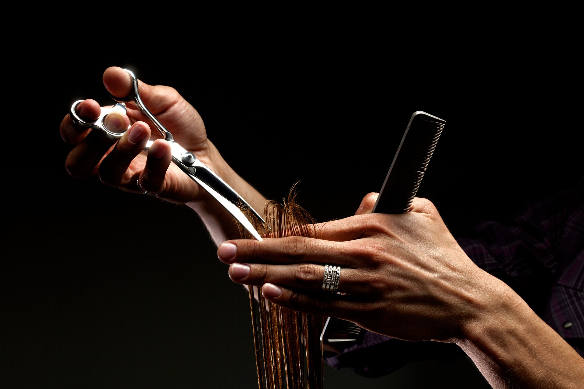 Hairdresser's hands hold scissors and a comb, cutting a lock of brown hair. Dark background.