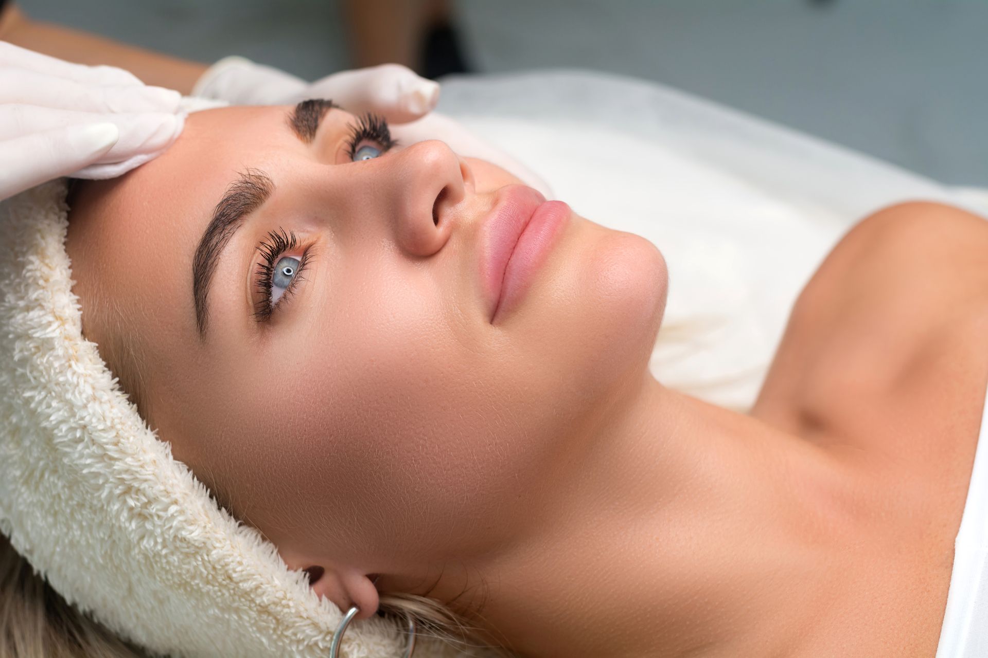 Woman with blue eyes and light skin receiving facial treatment, lying on a table.