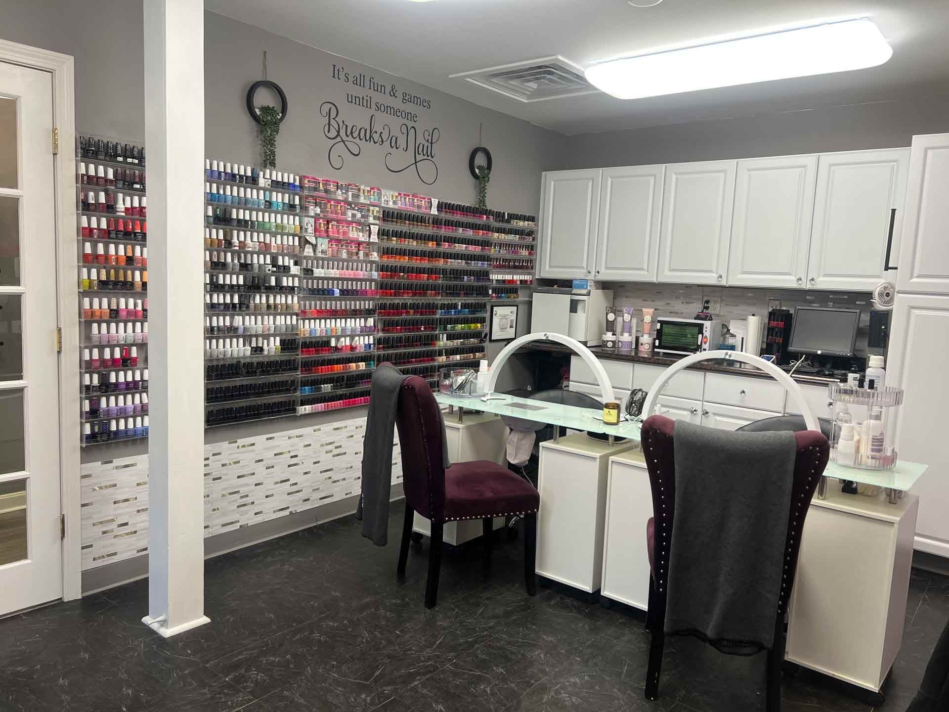 A nail salon station featuring two chairs, a white desk, and a wall filled with many bottles of colorful nail polish.