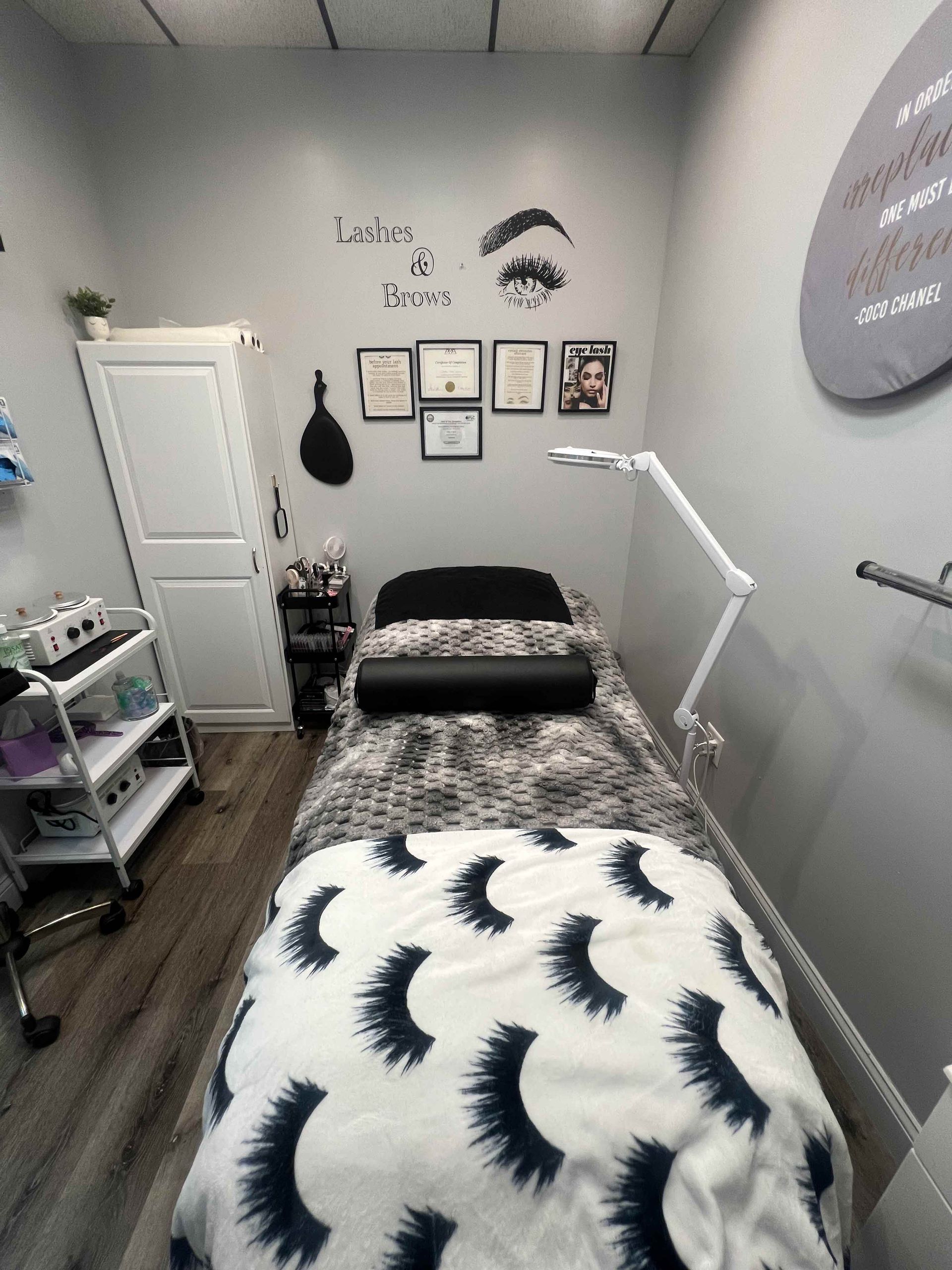 An eyelash extension studio features a treatment bed with a lash-pattern blanket, a tall lamp, and wall-mounted certificates.
