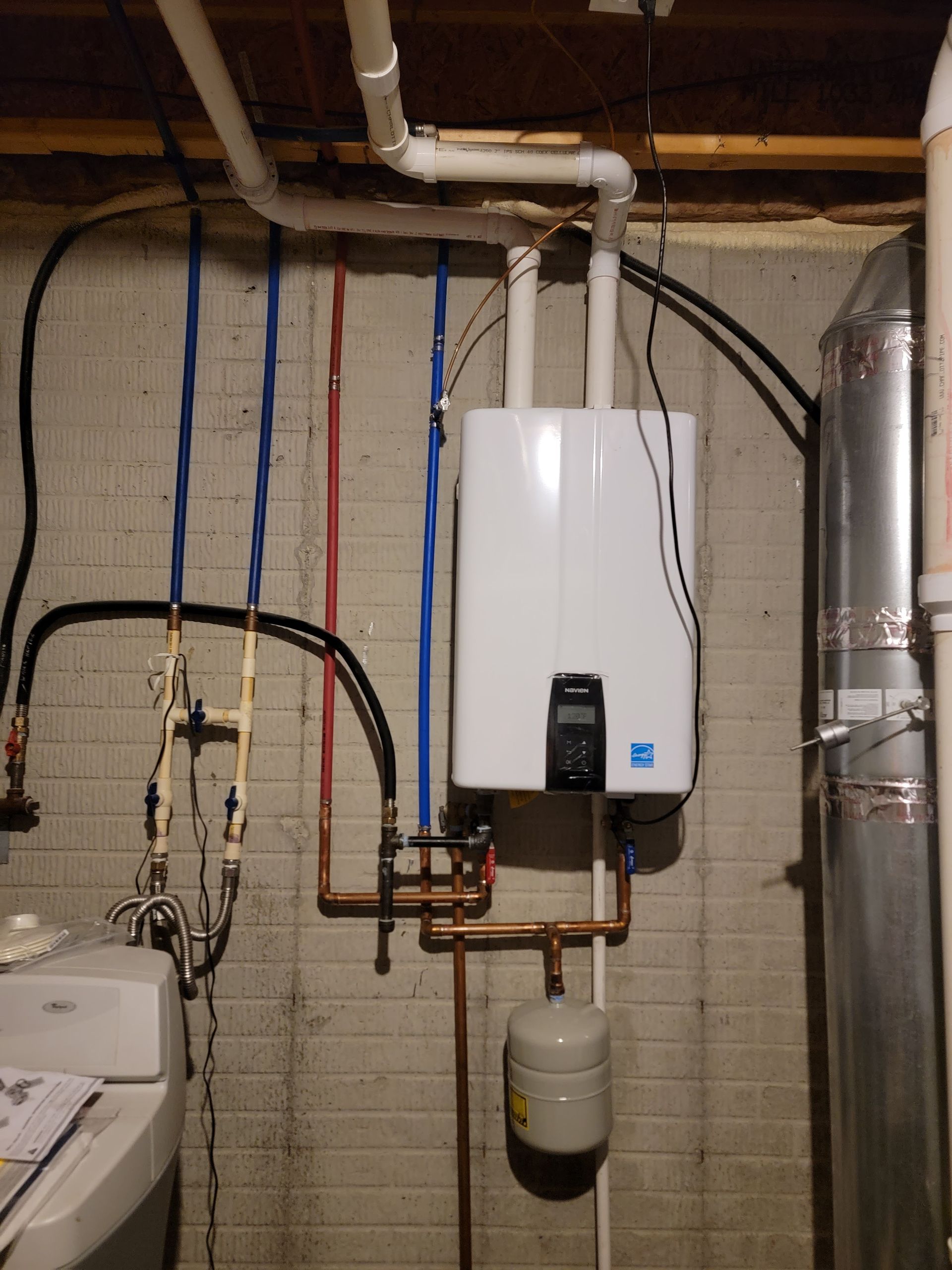 A white water heater is mounted to a brick wall in a basement
