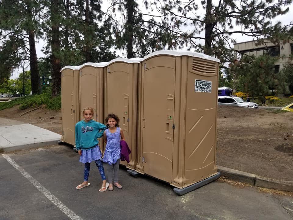 Two girls standing in front of four brown portable toilets in a parking lot.