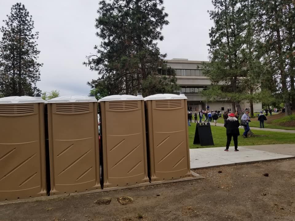 Four portable toilets in a row; people gather on a lawn in front of a building.