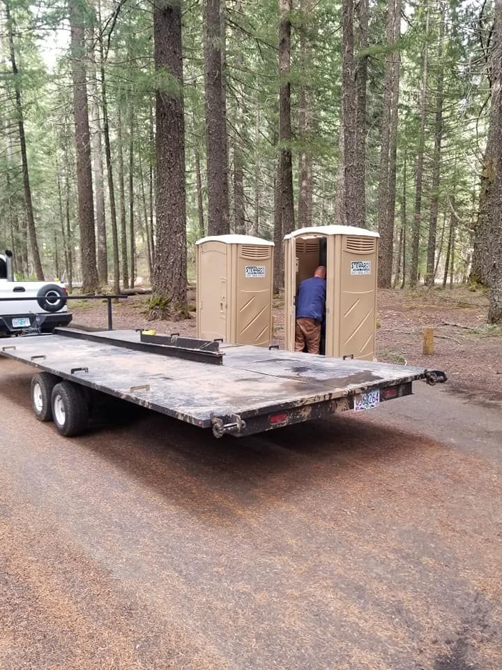 Two portable toilets on a trailer in a forest setting; person inside one.