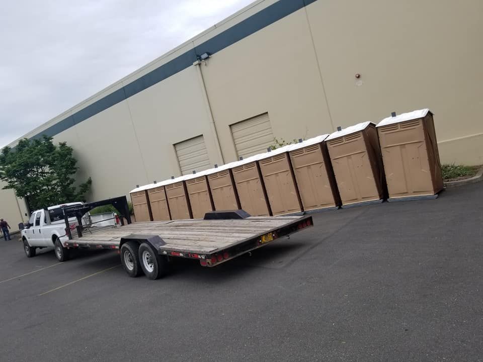 White truck towing an empty flatbed trailer. Row of brown portable toilets against a building.
