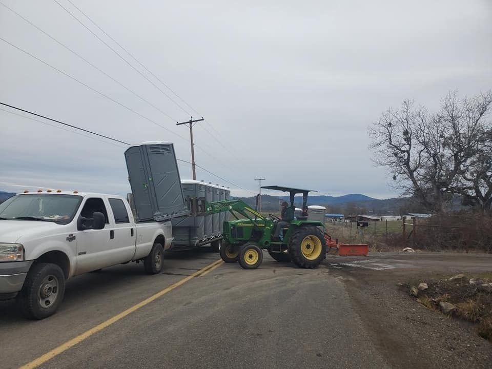 White truck and John Deere tractor towing a trailer of portable toilets on a road.