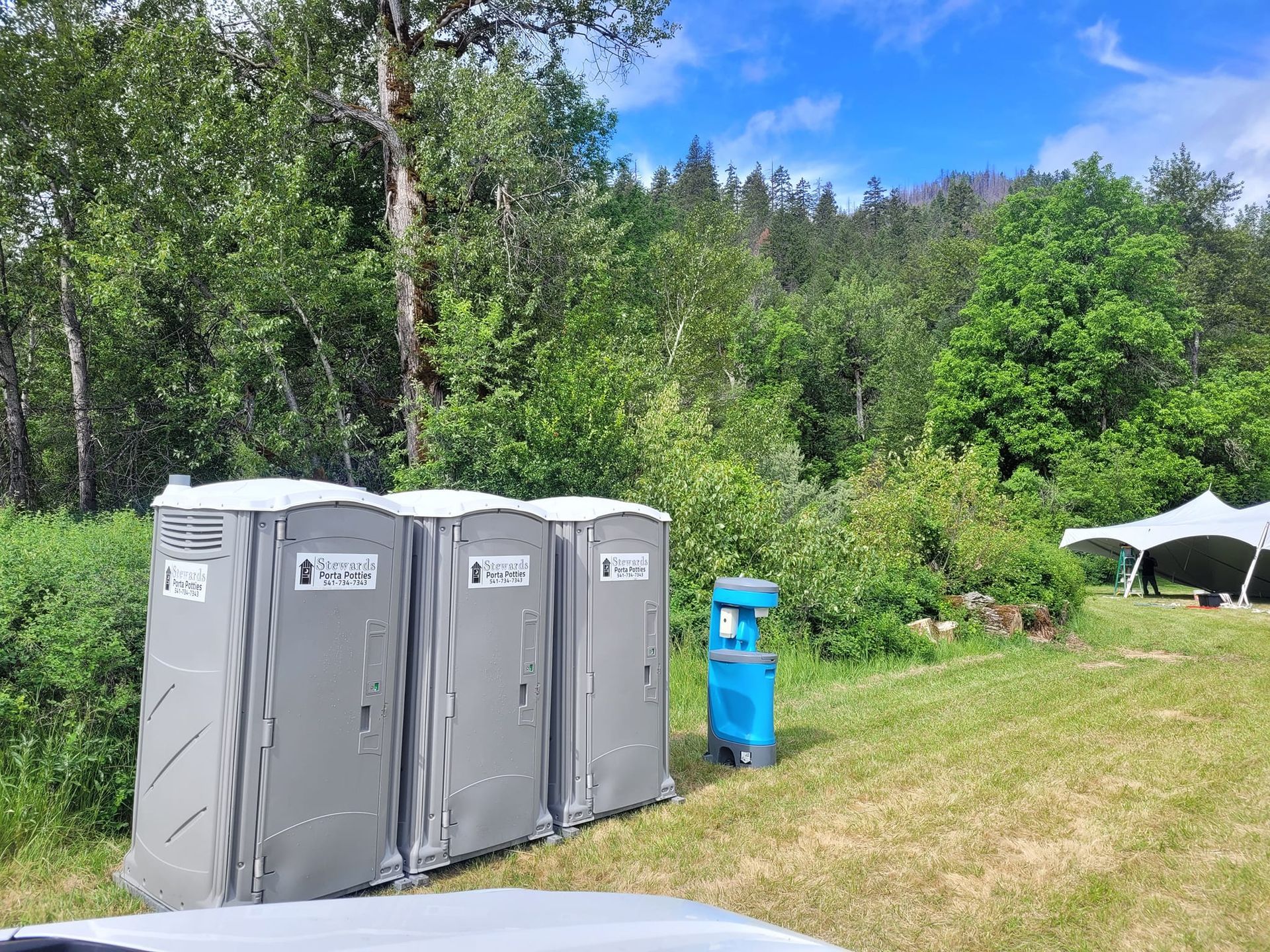 Three gray portable toilets and a blue handwashing station on a grassy area, trees in the background, a white tent visible.