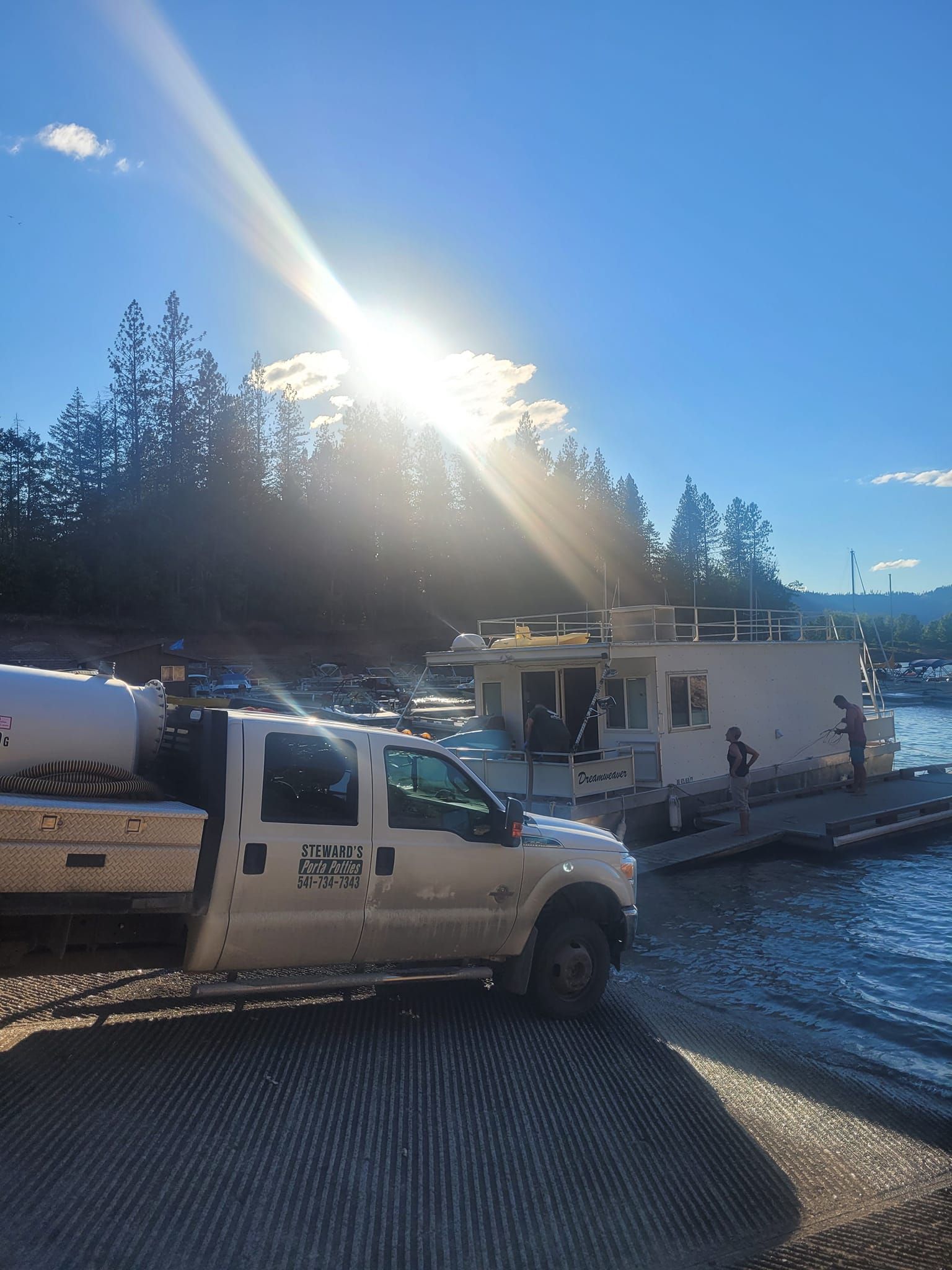 A white truck pulling a houseboat from a ramp into a lake, bright sun overhead.