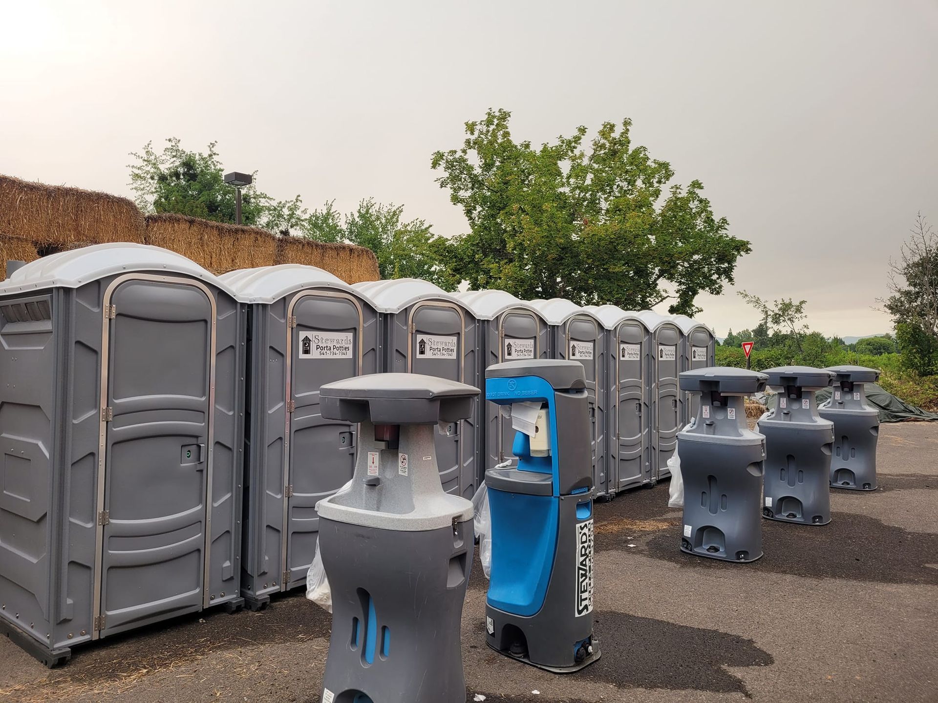 A row of portable toilets and water dispensers on a paved area, with trees and a cloudy sky in the background.