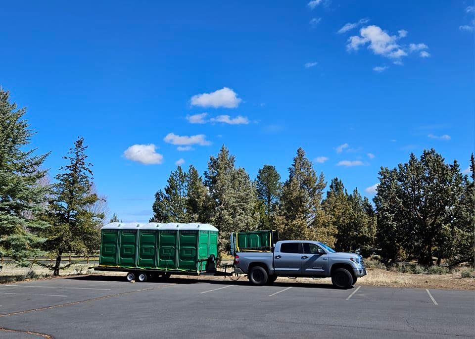 Truck towing a trailer with green portable toilets in a parking lot on a sunny day.