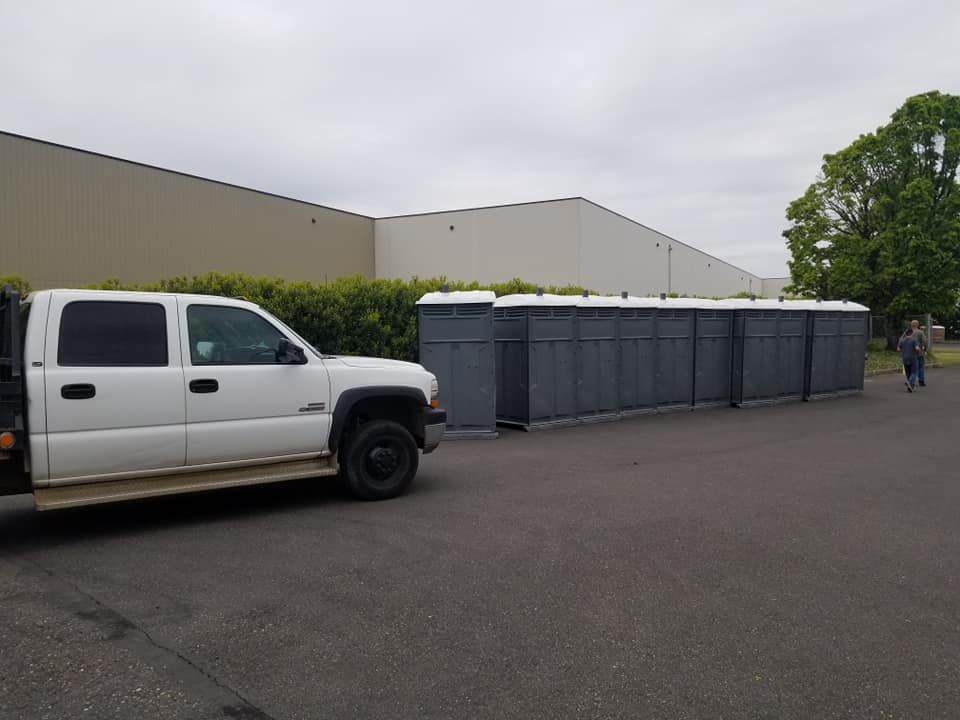 White truck and portable toilets in a parking lot. Person walks on the right. Beige building in the background.