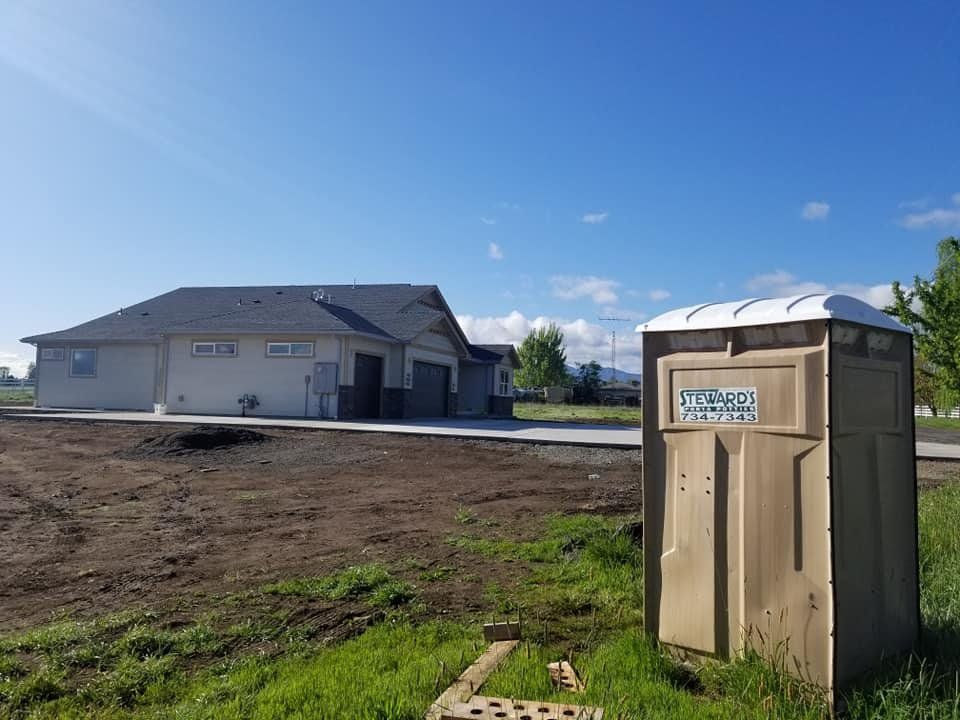 A tan portable toilet sits in front of a partially constructed building on a sunny day.