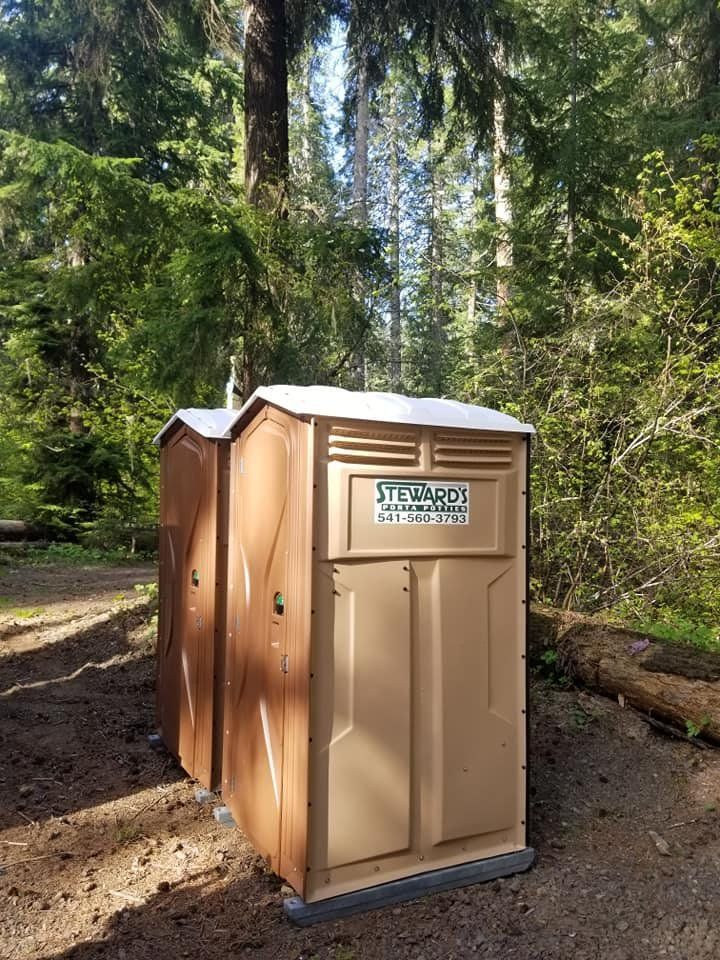 Two brown portable toilets in a wooded area with trees and sunlight.