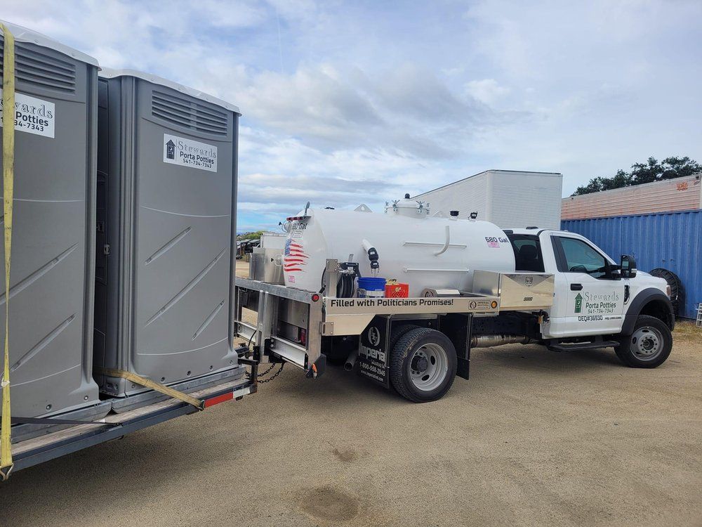 A white truck with a tank and portable toilets on a trailer, outdoors.