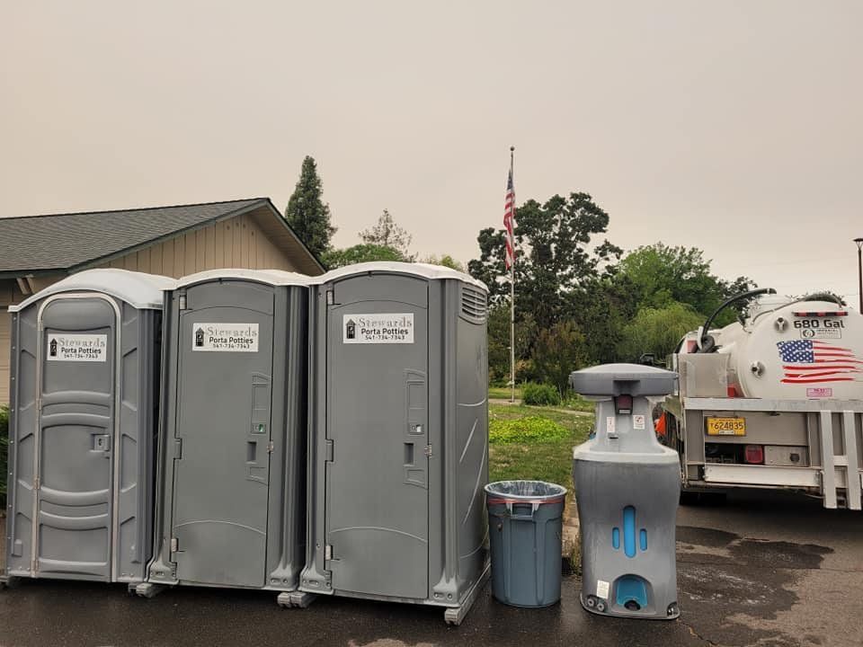 Three gray portable toilets and a handwashing station next to a parked truck with the American flag in the background.
