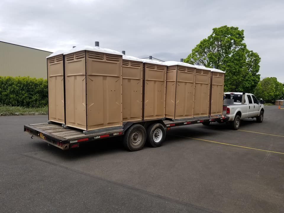 A truck towing a flatbed trailer loaded with multiple portable toilets on a paved lot.