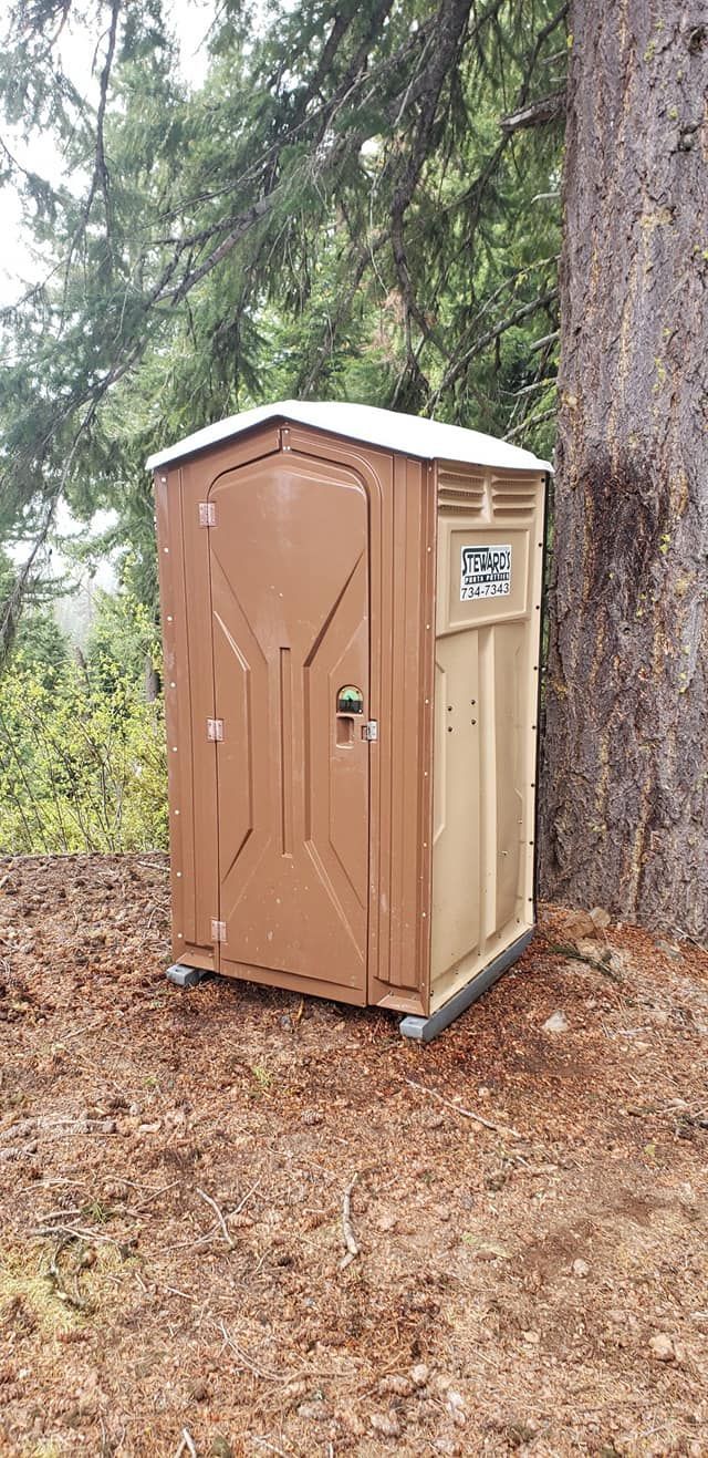 Brown portable toilet in a wooded area next to a large tree.