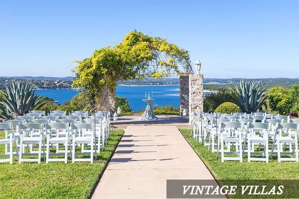 A row of white chairs are lined up in front of a stone archway overlooking a body of water.