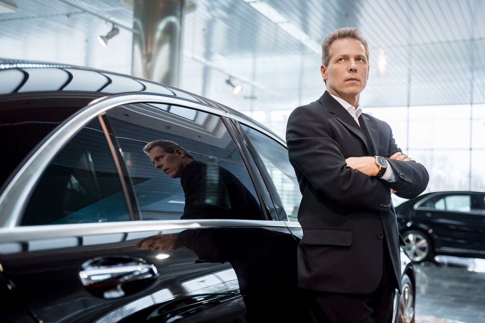Man in suit standing next to a black car in a dealership. Reflective windows and watch visible.