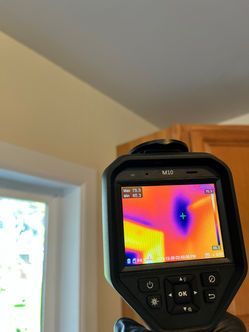 Thermal camera displaying heat signatures on a wall and ceiling near a window. Hot spot indicated in purple.