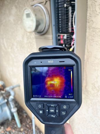 An infrared camera detecting heat from electrical panel. Display shows a bright red/yellow spot.
