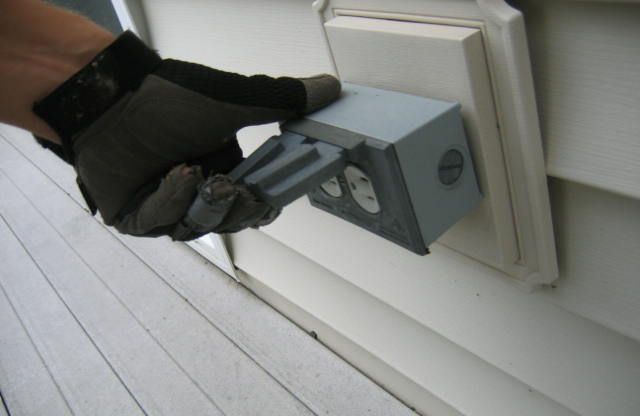Gloved hand opening a gray weatherproof electrical outlet cover on a white siding wall.