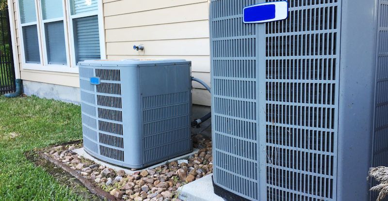 Two gray air conditioning units next to a house, sitting on rocks and concrete.