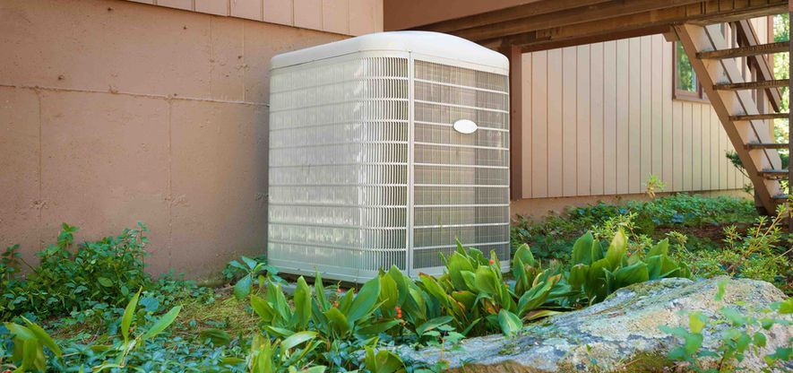 An air conditioning unit in a yard surrounded by plants and a rock, next to a wall and stairs.